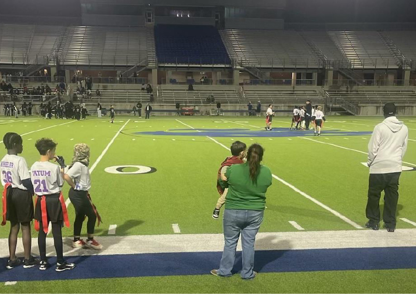 "Night-time youth flag football game at a stadium, with a woman holding a child and watching the game from the sidelines. Young players in jerseys stand nearby, and distant spectators are seated in the illuminated stands, creating a vibrant community sports environment.