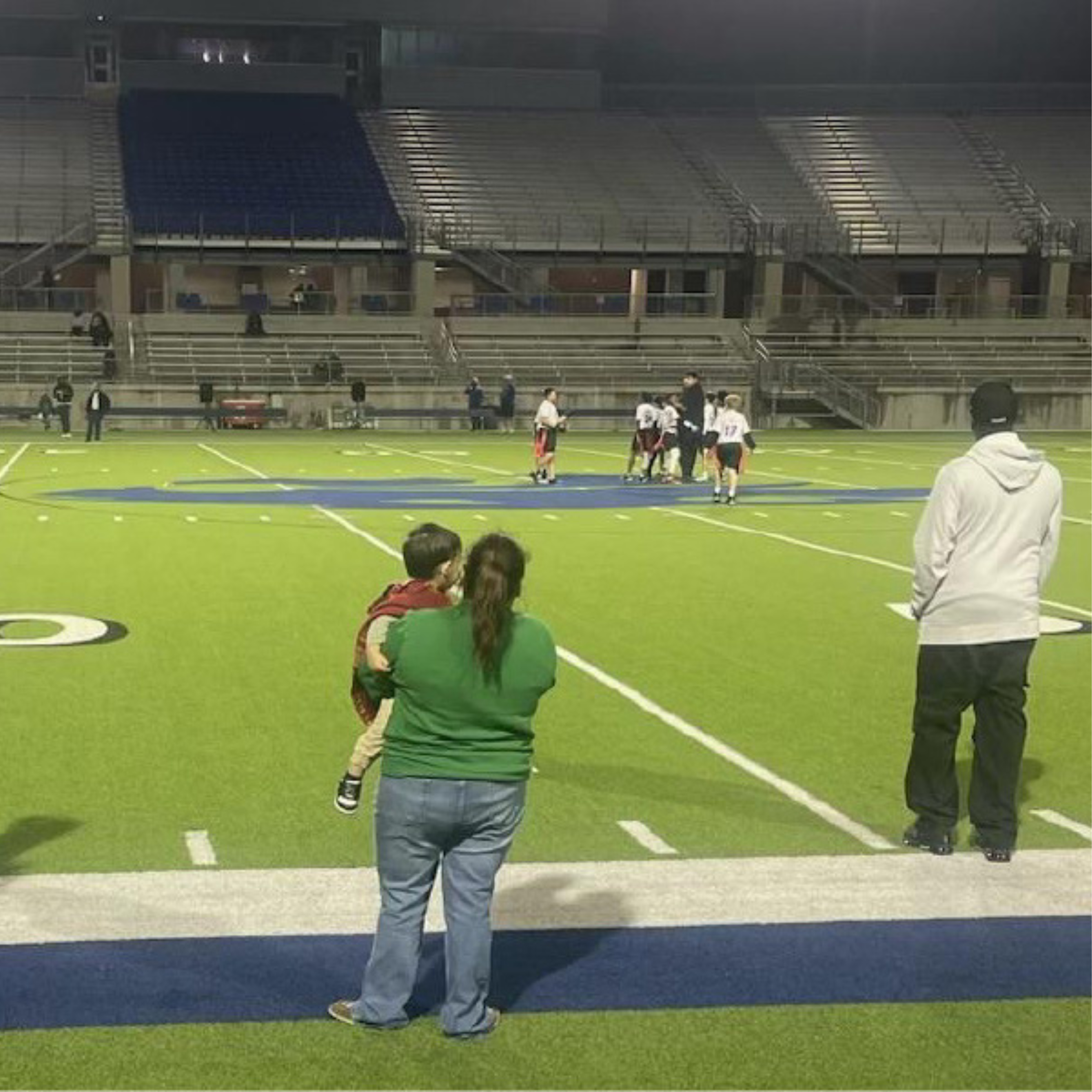ELQN web logos standard size (7) "Night-time youth flag football game at a stadium, with a woman holding a child and watching the game from the sidelines. Young players in jerseys stand nearby, and distant spectators are seated in the illuminated stands, creating a vibrant community sports environment.